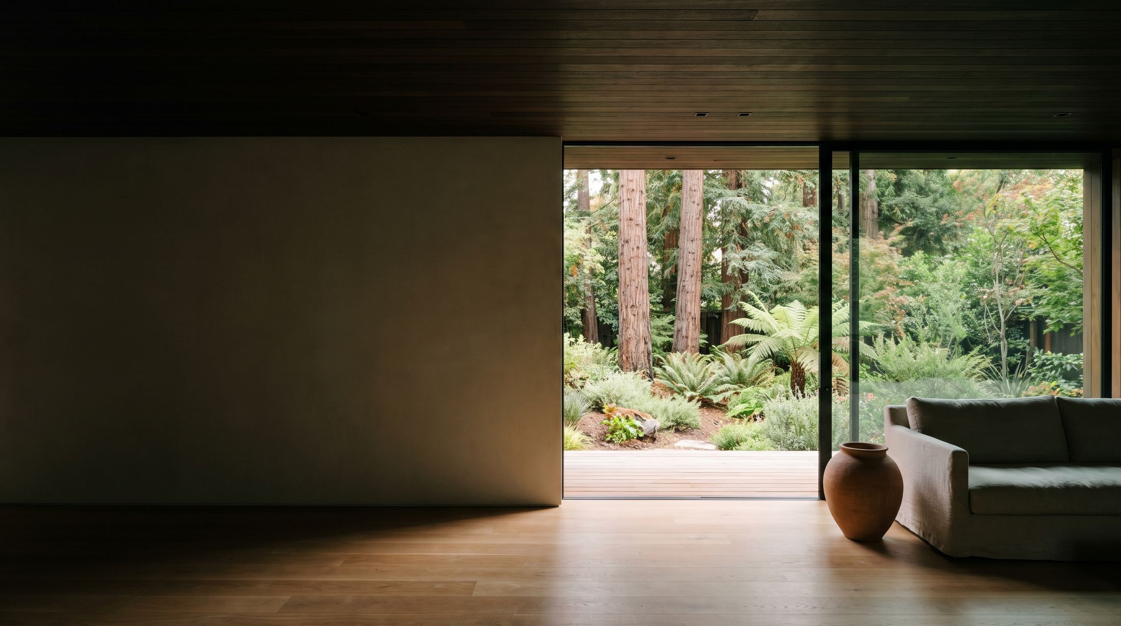 A Peninsula living room at mid-morning under a warm timber ceiling, opening through a full-height sliding glass wall onto a garden of native oaks and grasses.